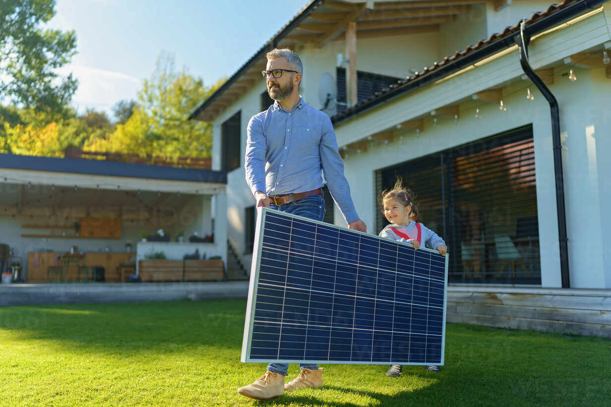 Father and young daughter carrying a large solar panel across a green backyard lawn, in front of a modern home with outdoor patio space and solar-friendly architecture.